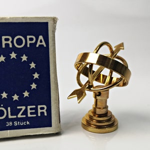 May include: A gold-colored metal globe with an arrow pointing upwards, on a base with a circular top. The globe is surrounded by a ring. The globe is next to a blue and white box with the text "EUROPA HÖLZER 38 Stück".