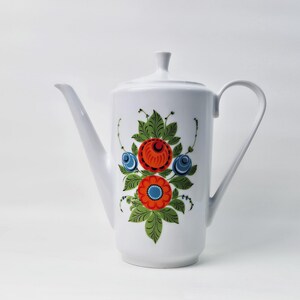 以下が含まれることがあります： 花柄の白い陶器製のティーポット。花は赤、オレンジ、青、緑です。ティーポットは注ぎ口と取っ手があります。