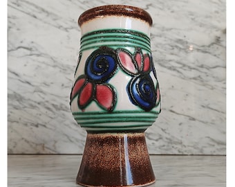 May include: A white ceramic vase with a brown base and green and blue floral designs. The vase has a flared top and a rounded base.