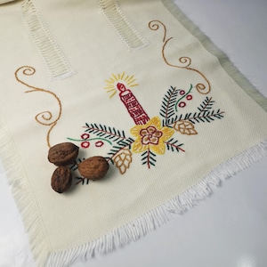 May include: White embroidered table runner featuring a red and yellow candle, green leaves, and brown nuts. The runner has a white fringed border.