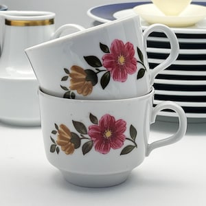 Op de afbeelding: Twee witte theekopjes op elkaar gestapeld, elk versierd met een bloemenpatroon met roze en bruine bloemen en groene bladeren. De kopjes hebben een afgeronde vorm met een oor. Ander wit en blauw serviesgoed staat op de achtergrond.