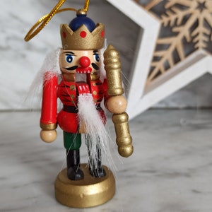 May include: A wooden nutcracker ornament with a red and gold uniform, a white beard, and a gold crown. The nutcracker is holding a gold and wooden staff.