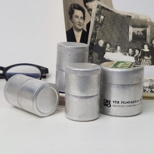 Könnte beinhalten: Eine Sammlung von Vintage-Filmdosen aus Aluminium in verschiedenen Größen, einige gestapelt, mit einigen alten Schwarz-Weiß-Fotografien und einer schwarzen Brille im Hintergrund. Die Dosen haben eine zylindrische Form.