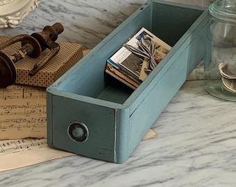 May include: A distressed, rectangular, light blue wooden drawer with a round knob. The drawer contains a stack of vintage postcards tied with a ribbon. The drawer is on a marble surface, with sheet music and other vintage items in the background.