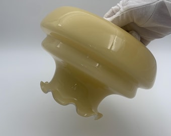 May include: A vintage, pale yellow glass lampshade with a ruffled edge. The shade has a rounded top and a flared, wavy bottom. The glass appears smooth and glossy. The lampshade is held by a gloved hand.