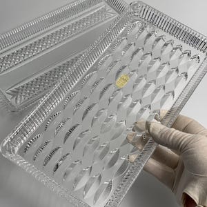 May include: Two clear rectangular trays with intricate geometric patterns. One tray features a diamond pattern, while the other has a series of curved, leaf-like designs. The trays are made of a transparent material, likely glass or crystal.