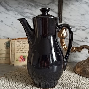 Könnte beinhalten: Eine schwarze Teekanne aus Keramik mit Deckel und Henkel. Die Teekanne steht auf einer weißen Spitzentischdecke.