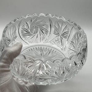 以下が含まれることがあります： スカラップトリムが付いた透明なカットガラスボウル。ボウルは、複雑なスターバーストと扇形のパターンを特徴としています。手袋をはめた手がガラスを持ち、装飾的なボウルの詳細な職人技とデザインを紹介しています。