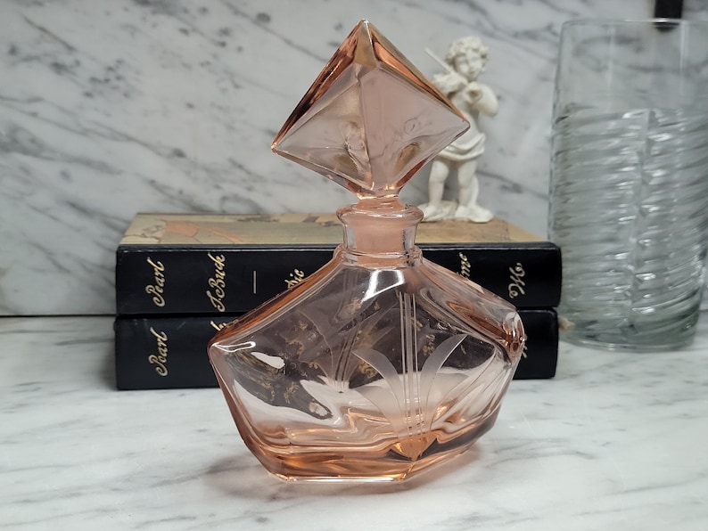 May include: A pink glass perfume bottle with a diamond-shaped stopper. The bottle has an etched design and a geometric shape.