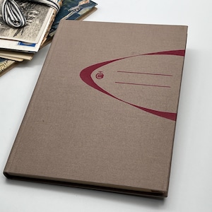 May include: A closed, brown book with a maroon design on the cover. The design includes a curved line and two horizontal lines for writing. Vintage postcards and a striped ribbon are visible in the background.