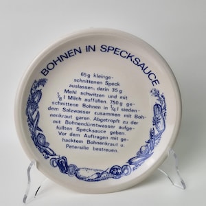 May include: A white ceramic plate with blue trim and a recipe for "Bohnen in Specksauce" printed on it. The recipe includes ingredients and instructions for making beans in bacon sauce.