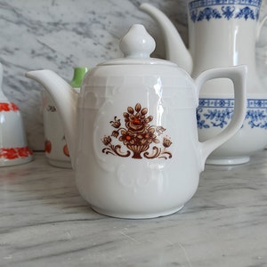 May include: White porcelain teapot with a floral design in shades of brown and orange. The teapot has a rounded body and a spout on the side. The handle is on the side of the teapot.