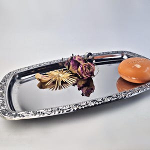 Könnte beinhalten: Ein rechteckiges, silberfarbenes Tablett mit verzierten Rändern, das ein orangefarbenes Seifenstück, getrocknete Rosen und einen dekorativen Strohstern enthält. Auf der Seife steht das Wort "Sandddon". Das Tablett spiegelt die Objekte wider.