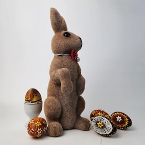 May include: A brown plush bunny rabbit with a red bow tie stands next to four decorated Easter eggs. The eggs are painted with intricate designs in shades of brown, yellow, and white.