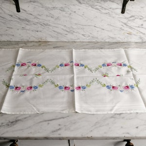 以下が含まれることがあります： ピンクとブルーの花が繊細な花柄で刺繍された白いテーブルランナー。デザインはランナーの長さに沿って繰り返されています。
