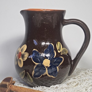 Può includere: Una brocca in ceramica marrone con un grande manico e un beccuccio. La brocca è decorata con fiori dipinti a mano nei toni del blu, giallo e bianco. L'interno della brocca è arancione. La brocca è appoggiata su un panno di pizzo bianco.
