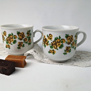 May include: Two white ceramic teacups with a floral pattern in shades of yellow, brown, and green. The cups have a simple design and are perfect for a classic tea party.