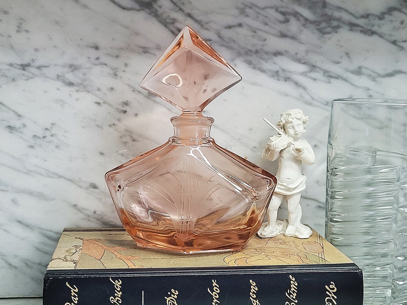 May include: A pink glass perfume bottle with a diamond-shaped stopper. The bottle has a geometric design and is sitting on a book.