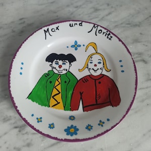 Peut inclure: Une assiette en céramique blanche avec un bord violet, représentant une illustration peinte à la main de deux clowns avec des nez rouges. L'assiette est décorée de fleurs bleues et jaunes et du texte "Max und Moritz".