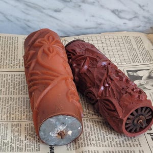May include: Two vintage paint rollers with intricate designs. The roller on the left has a floral pattern and is a light brown colour. The roller on the right has a more abstract design and is a dark red colour.