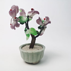 May include: A small, potted bonsai tree with purple glass flowers and green glass leaves. The tree is in a green ceramic pot with white sand.
