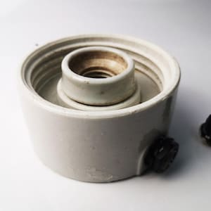 May include: White ceramic electrical outlet with a black plastic connector. The outlet has a threaded center hole and a raised lip around the edge.