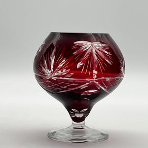 Könnte beinhalten: Ein rubinrotes Kristall-Brandyglas mit einem klaren, runden Fuß. Die Schale des Glases weist ein geschliffenes Design aus radialen Linien und stilisierten Blattformen auf. Das Glas ist zum Servieren von Brandy bestimmt.