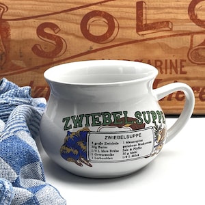 Può includere: Tazza da zuppa in ceramica bianca con manico, con una ricetta per Zwiebelsuppe (zuppa di cipolle) in tedesco. La tazza ha un'illustrazione blu e gialla ed è ambientata su uno sfondo di legno e un panno a scacchi blu e bianco.