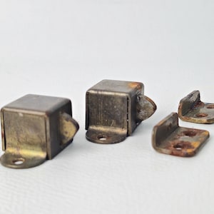 以下が含まれることがあります： ヴィンテージの金属製キャビネットラッチとキャッチのセット。セットには、丸みを帯びたキャッチが付いた2つの正方形のラッチと、2つのマッチングコーナーキャッチが含まれています。金属は変色し、古くなった外観で、錆のヒントがあります。