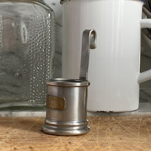 Könnte beinhalten: Ein kleiner, silberfarbener Messbecher mit Henkel, beschriftet mit "1/2 cl". Der Becher ist aus Metall und hat ein klassisches Design. Er steht auf einer Holzoberfläche, mit einem Glas und einer weißen Tasse im Hintergrund.