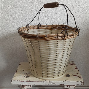 Könnte beinhalten: Ein weißer Weidenkorb mit einem braunen Holzgriff und einem Drahtgestell. Der Korb steht auf einem weißen Holzhocker.