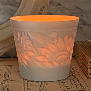 Può includere: Un portacandele bianco con un motivo a girasoli. Il portacandele è illuminato dall'interno, emettendo una calda luce. I girasoli sono in rilievo sul lato. Il portacandele è appoggiato su uno spartito musicale.
