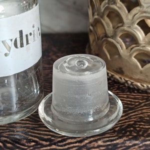 May include: A clear glass bottle stopper with a frosted finish. The bottle stopper has a wide base and a narrow neck. The label on the bottle reads "Hydric".