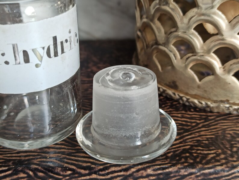 May include: A clear glass bottle stopper with a frosted finish. The bottle stopper has a wide base and a narrow neck. The label on the bottle reads "Hydric".