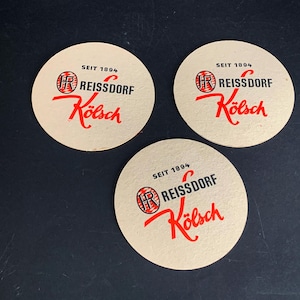 May include: Three beige round coasters with the text "SEIT 1894 REISSDORF Kölsch" in red and black. The coasters have a circular logo with the letters "JR" inside. The coasters are on a dark surface.
