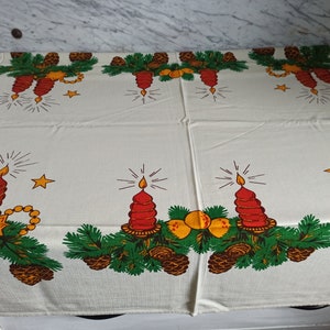 Op de afbeelding: Witte tafelkleed met een kerstthema met rode kaarsen, groene dennentakken, dennenappels en gouden sterren.