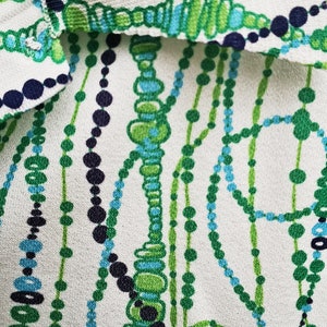 May include: White fabric with a repeating pattern of green, blue, and black beads. The beads are arranged in a geometric pattern.