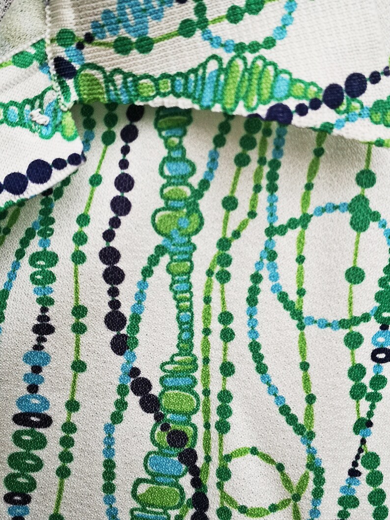 May include: White fabric with a repeating pattern of green, blue, and black beads. The beads are arranged in a geometric pattern.
