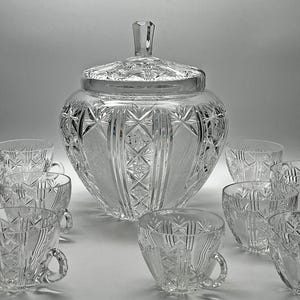 Könnte beinhalten: Ein Bowle-Set aus klarem Glas, bestehend aus einer großen Schüssel mit Deckel und mehreren passenden Tassen. Jedes Teil ist mit geometrischen Sternmustern und vertikalen Linien verziert, was eine facettierte und reflektierende Oberfläche erzeugt.