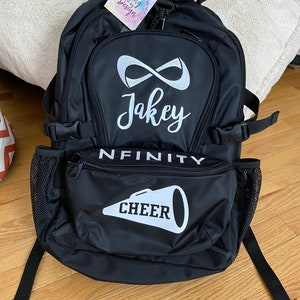 May include: Black backpack with a white infinity symbol and the name "Jakey" written in cursive. The backpack also has a white megaphone with the word "CHEER" written on it and the word "INFINITY" written in white letters.