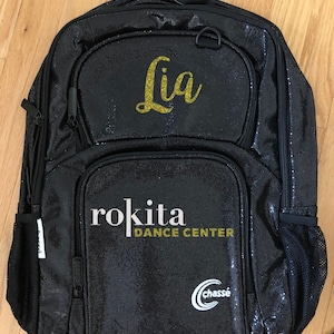 May include: Black glitter backpack with the name "Lia" in gold script. The backpack has multiple zippered compartments and a side mesh pocket. The text "roKita DANCE CENTER" and the "Chassé" logo are also visible.