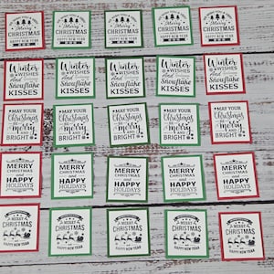 May include: A collection of twenty-five square paper tags featuring black printed holiday text and graphics on white backgrounds, each framed by a thin red or green border, including phrases like Merry Christmas and Winter Wishes.