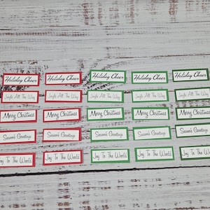 May include: A collection of Christmas-themed gift tags with various festive messages. The tags are rectangular, with text in a handwritten style. Some tags have red borders, while others have green borders. The messages include "Holiday Cheer," "Jingle All The Way," "Merry Christmas," "Season's Greetings," and "Joy To The World."