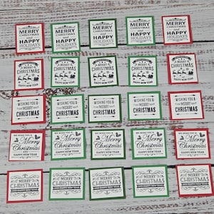 May include: A collection of twenty-five square paper tags arranged in five rows. Each tag features black printed holiday text and graphics, including phrases like Merry Christmas and Happy Holidays, framed by either red or green borders.