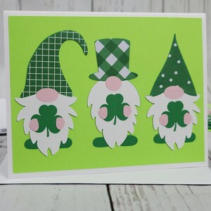 May include: A green card with three gnomes holding shamrocks. The gnomes are wearing green hats with white polka dots, green and white plaid, and a green hat with a white grid pattern. The card has a green background.