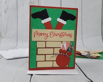 May include: A green Christmas card with a Santa Claus leg sticking out of a brick chimney. The card says "Merry Christmas" in red letters.