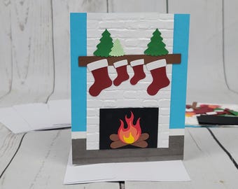 May include: A white brick fireplace with a brown mantel and stockings hanging from it. There are two green trees on the mantel and a red and yellow fire in the fireplace.