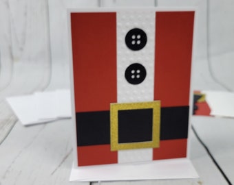 May include: A red and white Christmas card with a Santa Claus design. The card features a red suit, a white belt with a gold buckle, and two black buttons.
