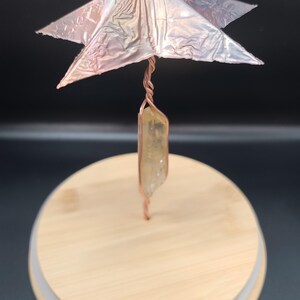 May include: A handcrafted decorative piece featuring a copper wire structure with a star-shaped top and a crystal pendant. The piece is set on a wooden lid, creating a unique and artistic display.