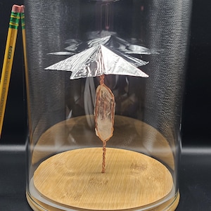 Può includere: Una campana di vetro trasparente con base in legno ospita una scultura di cristallo. La scultura presenta un cristallo trasparente sospeso con filo di rame, sormontato da un ombrello in lamina d'argento. Due matite sono visibili a sinistra.